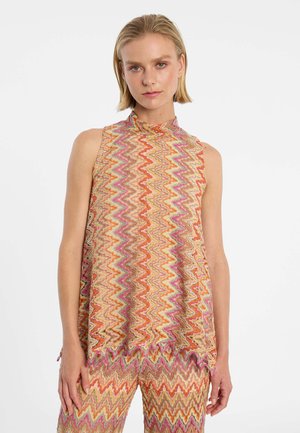 Frau trägt ärmelloses Oberteil mit Zickzackmuster und passende Hose in Orangetönen, Pink und Beige, steht vor weißem Hintergrund.