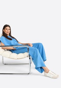 L'ensemble azzurro chiaro presenta una maglia dal taglio rilassato e pantaloni a gamba larga. Il modello è seduto su una poltrona morbida e testurizzata con dettagli in cromo. Le sneakers bianche completano l'outfit.