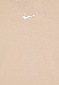 Bež majica iz bombaža s kratkimi rokavi, ki ima krožni izrez in belo vezeno Nike ikono na zgornjem levem prsnem delu. Mehka tekstura, enostaven dizajn.