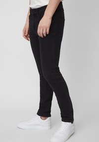Jeans de mezclilla negra con un corte ajustado, que cuentan con una bragueta de botón y cremallera, cinco bolsillos y detalles de costura. Combinados con zapatillas blancas.