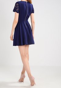 Navy blue dress with short lace sleeves, flared skirt, and back zipper detail. Paired with nude high-heeled shoes. Smooth texture.
