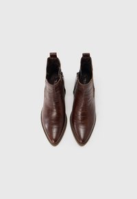 Para brązowych skórzanych botków za kostkę z szpiczastymi noskami i zamkami bocznymi, widzianych z góry na białym tle.