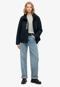 Granatowa kurtka z detalem logo, z zapięciem na zamek, dwoma kieszeniami na piersi i czarnymi akcentami. Zestawiona z jasnoniebieskimi spodniami o szerokich nogawkach i czarnymi butami.