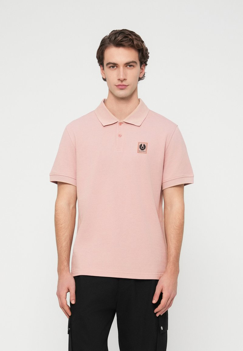 Polo rose pâle en coton, avec col, patte à trois boutons et petite étiquette logo sur le côté gauche de la poitrine.