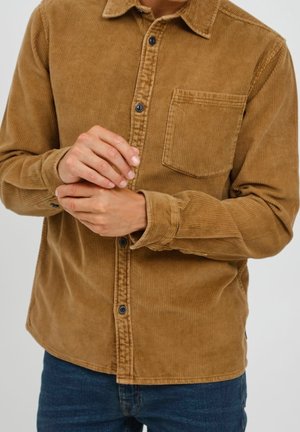 Mężczyzna w brązowej, sztruksowej koszuli zapinanej na guziki z kieszenią na piersi, poprawiający lewy mankiet, ubrany w niebieskie dżinsy na jednolitym tle.