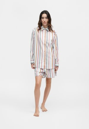 Γυναίκα που στέκεται ξυπόλητη, φορά μακρυμάνικο, ριγέ πουκάμισο με κουμπιά και αντίστοιχο ριγέ σορτς μπροστά σε απλό λευκό φόντο.