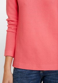 Koronkowa, prążkowana bluzka z długim rękawem w kolorze koralowym z okrągłym dekoltem, charakteryzująca się luźnym krojem i podwiniętymi mankietami, w zestawie z ciemnoniebieskimi jeansami o fakturze.