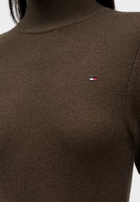 Κοντινό πλάνο από σκούρο καφέ πουλόβερ με λαιμόκοψη, φτιαγμένο από πλέξη, με μικρό κόκκινο, λευκό και μπλε ορθογώνιο λογότυπο στο στήθος.