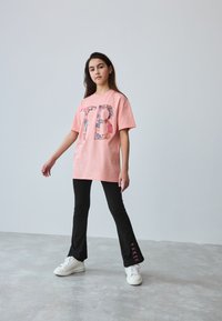 Lyserød oversized t-shirt med palietbesat "TB"-grafik, kombineret med tætsiddende sorte vide bukser med teksten "BAKER" i lyserød på kanten, og hvide sneakers.
