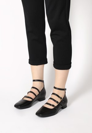 Schwarze Lederflachschuhe mit spitzem Zeh und vier dünnen Riemen, die goldene Schnallen haben. Der Absatz ist niedrig und quadratisch geformt.