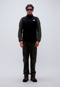 Άνδρας που φοράει σκούφο εξωτερικό γιλέκο, μακρυμάνικο πουκάμισο, παντελόνια, πεζοπορικά παπούτσια και γυαλιά ηλίου με μοβ απόχρωση, στέκεται μπροστά σε απλό φόντο.