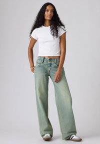 Γυναίκα με μακριά σγουρά μαλλιά που φοράει λευκό κοντό T-shirt, φαρδιά ξεθωριασμένα πράσινα τζιν και λευκά αθλητικά παπούτσια με μαύρες ρίγες.