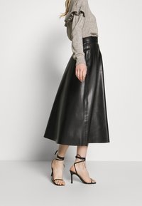 Femme portant un pull beige à volants, une jupe midi en cuir noir taille haute et des sandales à talons aiguilles noires, se tenant de profil.