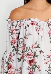 Bílý top bez ramínek s květinovým vzorem v růžových a zelených tónech, s volánkovým výstřihem a volnými rukávy. Měkká, lehká tkanina.