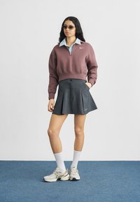 Γυναίκα με γυαλιά ηλίου, μοβ φούτερ με τέταρτο φερμουάρ, μπλε πουκάμισο με γιακά, πλισέ ανθρακί φούστα, άσπρες κάλτσες και μπεζ αθλητικά παπούτσια Nike.