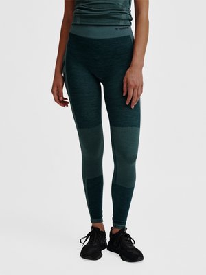 Kvinde iført mørkegrønne strukturerede leggings og sorte sportssko, der står mod en ensfarvet lys baggrund.