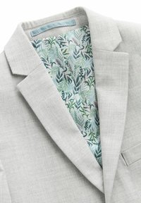Blazer gris clair avec revers crantés, montrant une doublure à motif de feuilles tropicales dans des teintes de vert et de bleu. Texture douce, coupe ajustée.