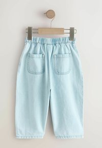 Pantaloni din denim albastru deschis cu talie elastică, două buzunare cusute în spate și tivuri răsucite, agățate pe un umeraș din lemn și metal.