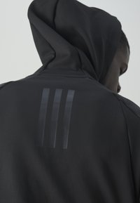 Personne portant une veste à capuche noire montrant trois bandes verticales brillantes au dos sur un fond clair uni.
