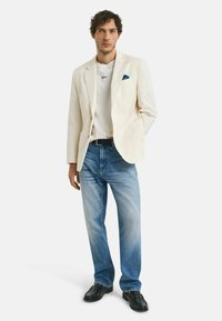 Blazer en lin blanc sur un t-shirt blanc avec "Good" brodé, jeans déchirés bleu clair et chaussures en cuir noir, les mains dans les poches.