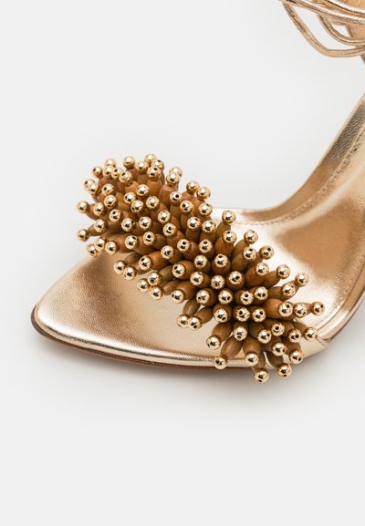 Scarpa con tacco alto dorata e metallizzata, con punta affusolata e un insieme di accentuazioni scultoree dorate nella parte anteriore. Texture liscia e design elegante.