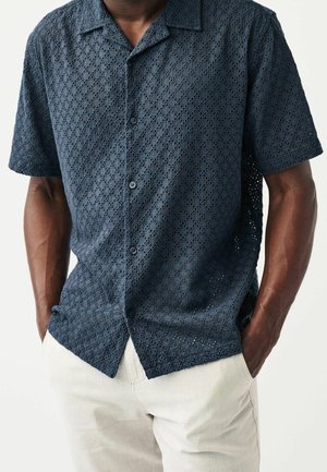 Mann trägt ein dunkelblaues Kurzarmhemd mit gemusterter Struktur und hellbeiger Hose, Hände in den Taschen.