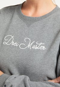 Grijze sweatshirt met geribbelde halslijn en manchetten. Bevat opvallende witte geborduurde tekst "Drei Master" op de borst. Zacht materiaal.