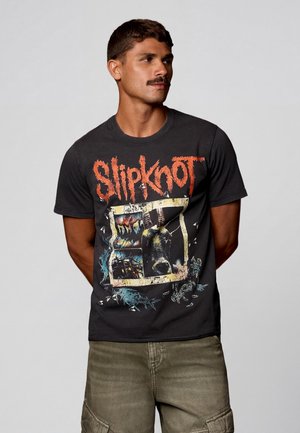 Bajszos férfi fekete Slipknot zenekaros pólóban, piros logóval és grafikával, zöld cargo rövidnadrággal, egyszínű háttér előtt.