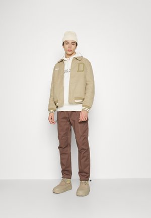 Beige Varsityjacke mit weißen Streifen, braunen Cargo-Hosen, cremefarbenem Hoodie und off-white Mütze. Merkmale den Buchstaben "D" auf der Jacke.