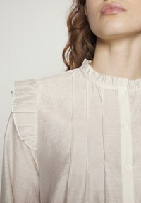Blusa beige chiaro con colletto arricciato, pieghe verticali, trama sottile e chiusura frontale con bottoni.
