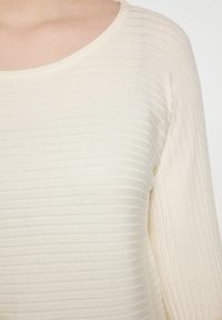 Maglione a coste di un beige chiaro con scollatura tonda, caratterizzato da un motivo testurizzato e maniche lunghe, realizzato in un tessuto morbido e leggero.