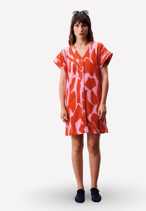 Femme debout portant une robe boutonnée à manches courtes avec un imprimé abstrait rouge et rose, des baskets noires et des lunettes de soleil sur la tête.