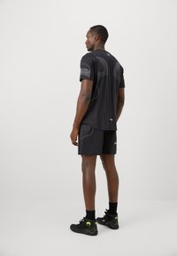 Mann iført svart sportstøy og svarte høye sneakers med neongrønne logoer, stående med ryggen til mot en ensfarget hvit bakgrunn.