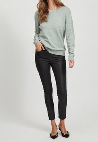 Světlé šedé pletené svetr s žebrovanými manžetami, v kombinaci s černými lesklými skinny kalhotami a černými muly s špičatou špičkou, které mají zlaté detaily.