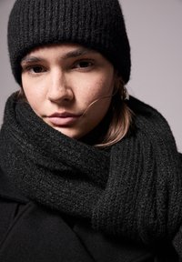 Beanie e sciarpa neri a costine, materiale texturizzato. Primo piano con pelle liscia e capelli morbidi, su uno sfondo tenue.