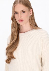 Pulover tricotat de culoare crem, cu guler rotund și textură ribbată, asortat cu păr ondulat și cercei mici cu cerc.