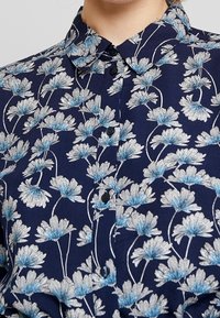 Marinblå skjorta med knappar som har ett blommönster i vitt och ljusblått, med spetsig krage och svarta knappar. Texturerat tyg.