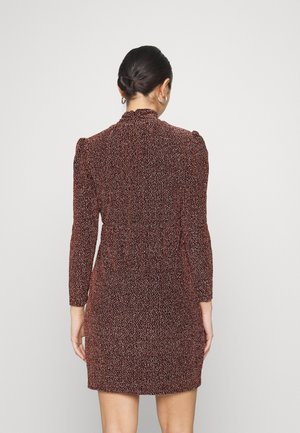 Femme aux cheveux foncés en chignon, portant des boucles d'oreilles créoles argentées et une mini-robe texturée à manches longues, debout de dos sur un fond uni.