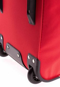 Valise en tissu rouge avec des accents en plastique noir, coins arrondis, deux roues et quincaillerie métallique en bas pour la durabilité.