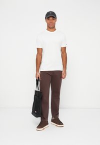 Camiseta blanca de manga corta, pantalones marrones, zapatillas oscuras y una gorra de béisbol negra. Sosteniendo una bolsa de mano negra. Atuendo simple y casual.
