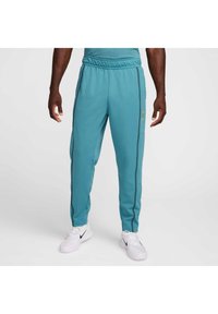 Turkise treningsbukser med elastisk midje, sidestriper og glidelåslommer. Laget av et glatt materiale. Kombinert med hvite joggesko.