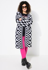Un long cardigan à motif carreaux noir et blanc, assorti à un t-shirt graphique noir et un pantalon rose vif. Le mannequin porte des lunettes de soleil noires surdimensionnées et des bottes noires.