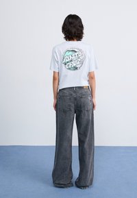 Lys blå t-skjorte med et sirkulært "Santa Cruz"-grafikk i grønt og hvitt, kombinert med løse, mørkegrå jeans. Enkel, uformell design.