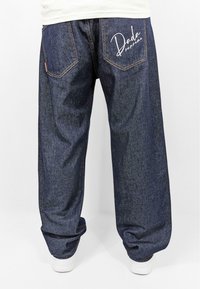 Denim trousers with a loose fit, dark blue color, stitched pockets, and white embroidered text "Dada" on the right rear pocket.