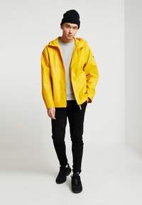 Giacca impermeabile gialla con cappuccio, abbinata a una felpa grigia e pantaloni neri slim-fit. Sneakers nere completano l'outfit.