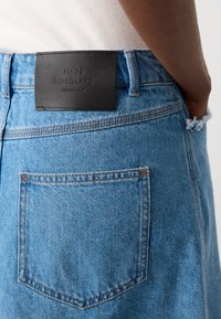 Blå denimskjørt med et svart lærmerke. Har en baklomme med oransje søm og opprevet kant nederst.