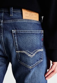 Jeans de mezclilla azul con una etiqueta de cuero en la cintura, que presentan costuras contrastantes en color marrón y un patrón de zigzag distintivo en el bolsillo trasero.