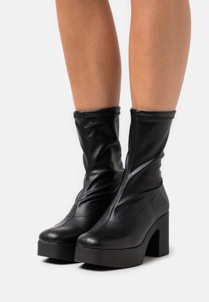 Černé kotníkové boty s výraznou siluetou, vyrobené z hladké kůže. Mají mohutný vysoký podpatek a jemné švové detaily.