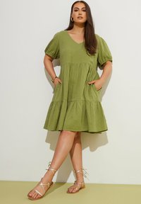 Olivengrünes, knielanges Kleid mit Puffärmeln, V-Ausschnitt, gestuftem Design und vorderen Taschen. Kombiniert mit sandfarbenen, schlichten Sandalen.