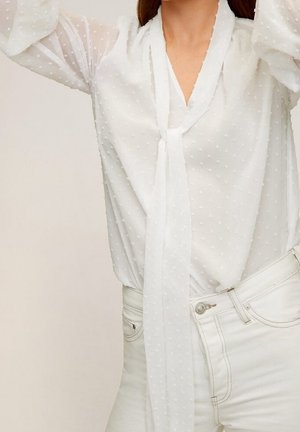 Kvinde iført en hvid, gennemsigtig prikket bluse med et slips om halsen og hvide højtaljede jeans, armene løftet over hovedet.